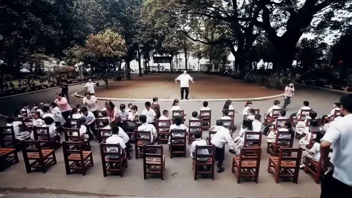 Students seated on rows of brown recycled-plastic school chairs in a tree-lined schoolyard