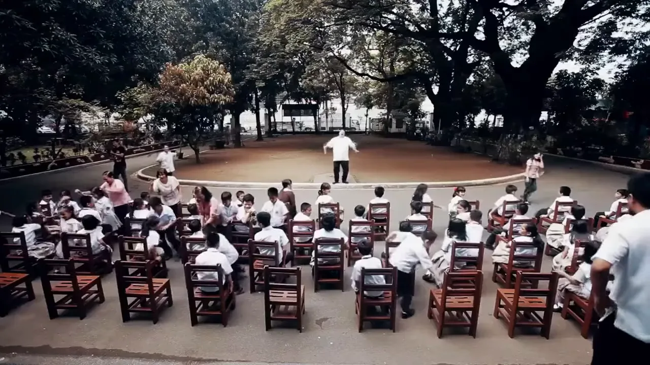 Students seated on rows of brown recycled-plastic school chairs in a tree-lined schoolyard